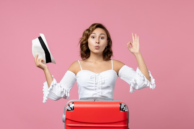 Front view of young woman holding hat and preparing for trip with red bag on pink wall