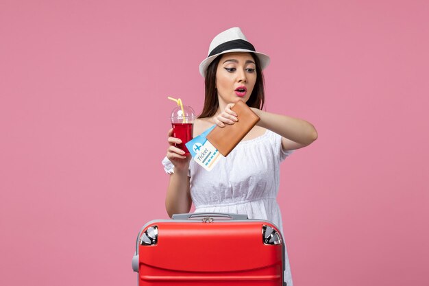 Вид спереди молодая женщина, держащая сок и билеты в отпуске на розовой стене, путешествие, летняя женщина