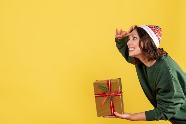 Front view of young woman holding little christmas present on yellow wall