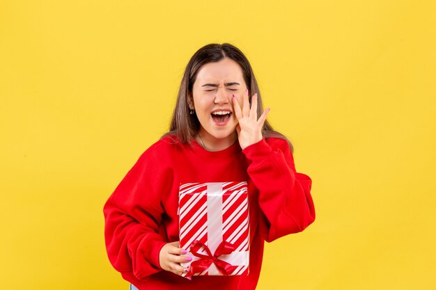 Front view of young woman holding little gift and screaming on yellow wall