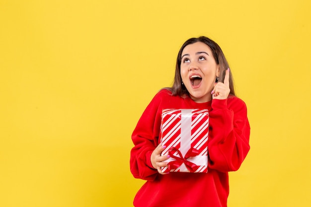 Front view of young woman holding little gift on the yellow wall