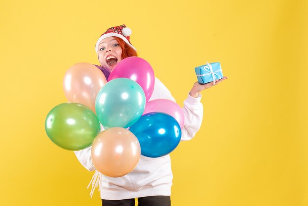 Front view of young woman holding little present and colorful balloons on a yellow wall