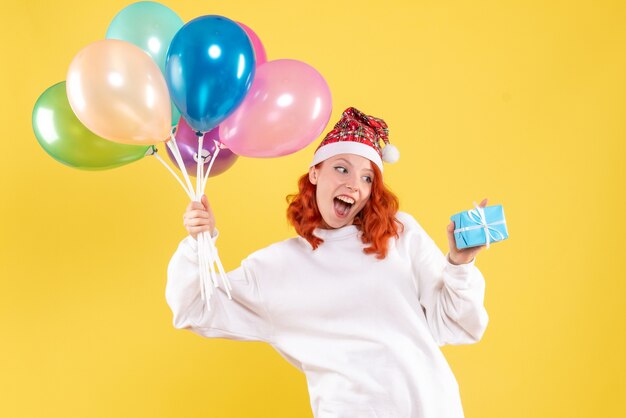 Front view of young woman holding little present and colorful balloons on yellow wall