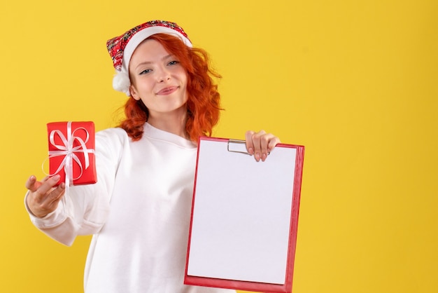 Front view of young woman holding little xmas present and file note on yellow wall