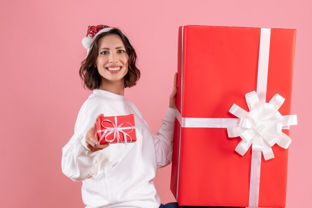 Вид спереди молодой женщины, держащей маленький рождественский подарок с огромным на розовой стене