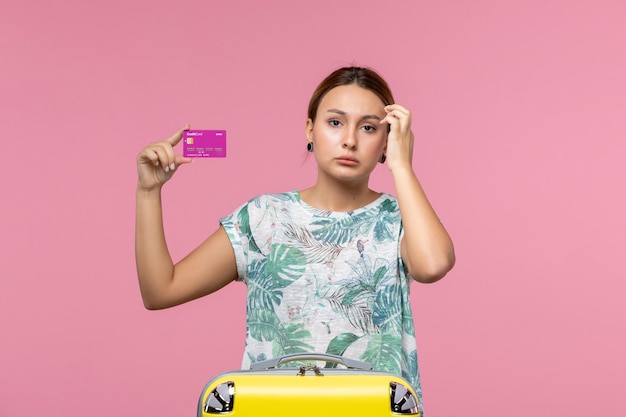 Front view young woman holding purple bank card on vacation on pink wall color voyage vacation summer woman
