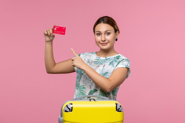 Вид спереди молодая женщина, держащая красную банковскую карту в отпуске на розовой стене, женщина вояж на самолете, рейс, отдых, отдых