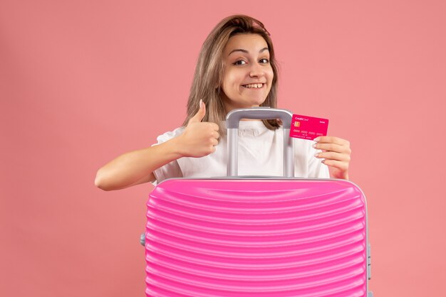 Front view young woman holding ticket giving thumbs up behind pink suitcase