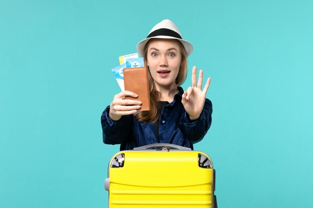 Вид спереди молодая женщина, держащая билеты и готовящаяся к поездке на голубом фоне, путешествие на море, отпуск, путешествие на самолете