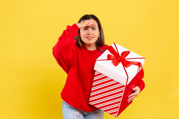 Front view of young woman holding xmas gift in box looking on yellow wall