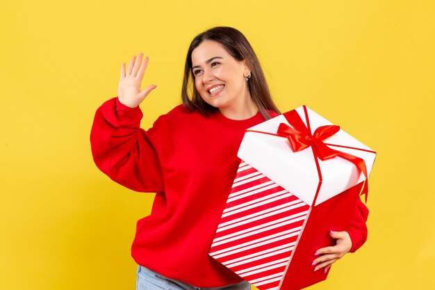 Front view of young woman holding xmas gift in box on the yellow wall
