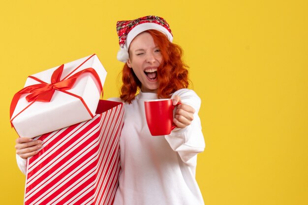 黄色の壁にクリスマスプレゼントとお茶を保持している若い女性の正面図