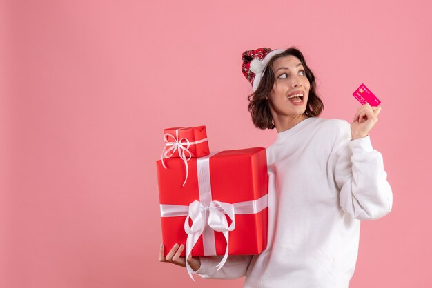 Front view of young woman holding xmas presents and bank card on pink wall