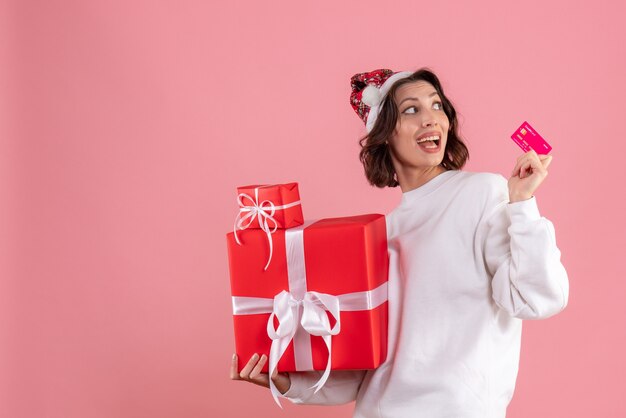 Вид спереди молодой женщины, держащей рождественские подарки и банковскую карту на розовой стене
