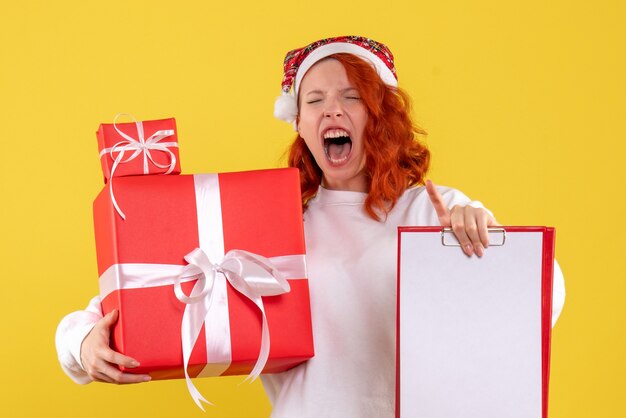 Вид спереди молодой женщины, держащей рождественские подарки и файловой заметки на желтой стене