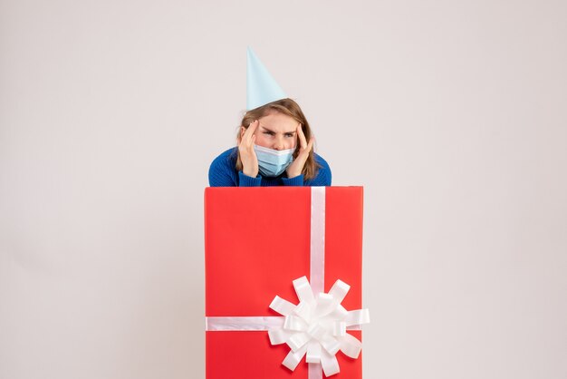 Front view of young woman inside present box in mask on the white wall