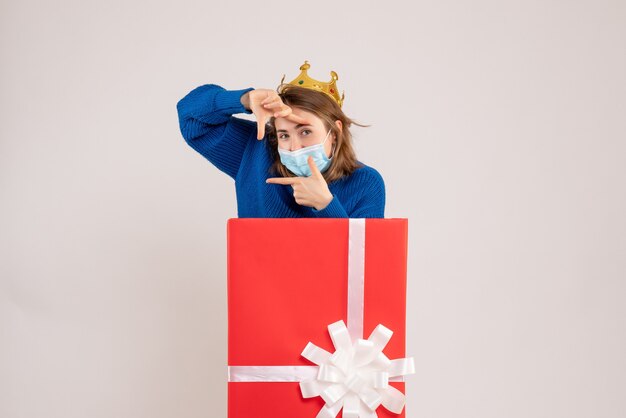 Front view of young woman inside present box in mask on white wall