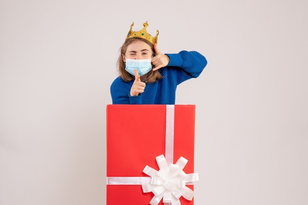 Front view of young woman inside present box in mask on white wall