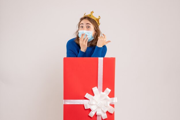 Front view of young woman inside present box in sterile mask on the white wall