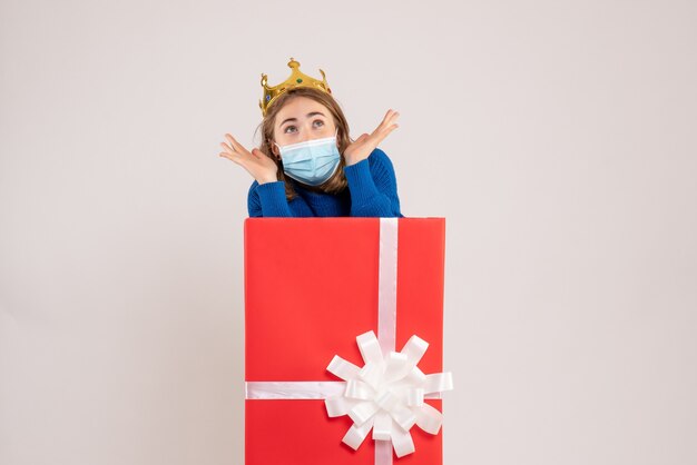 Front view of young woman inside present box in sterile mask on white wall