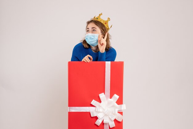 Front view of young woman inside present box in sterile mask on white wall