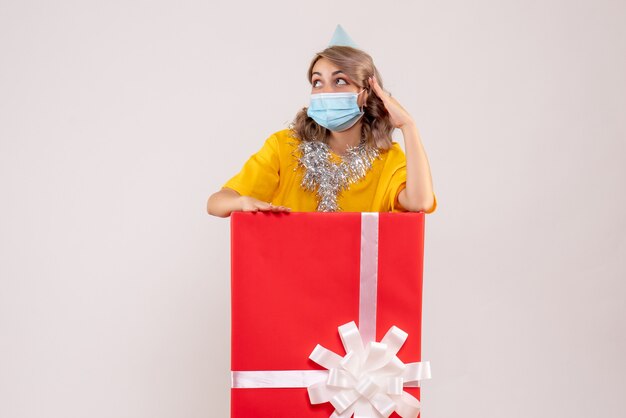 Front view of young woman inside red present box in mask on white wall