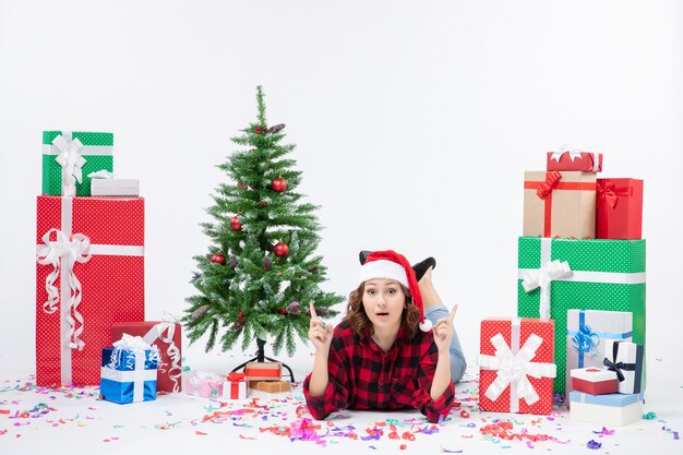 Front view of young woman laying around christmas presents and little holiday tree on white wall