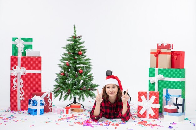 Front view of young woman laying around christmas presents and little holiday tree on white wall