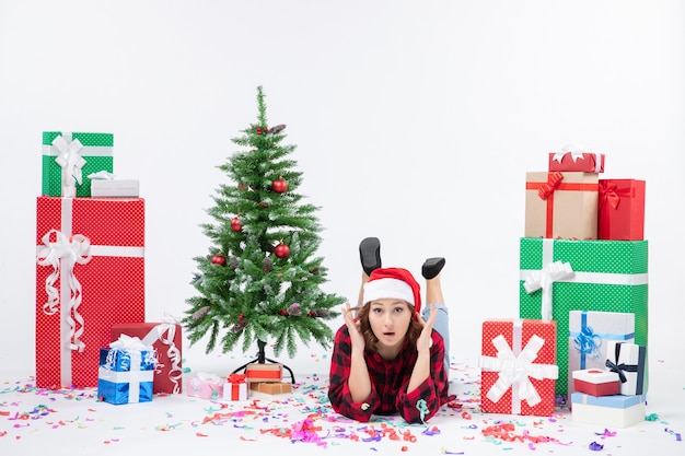 Front view of young woman laying around christmas presents and little holiday tree on white wall