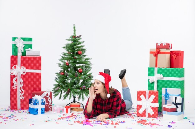 Front view of young woman laying around christmas presents and little holiday tree on white wall