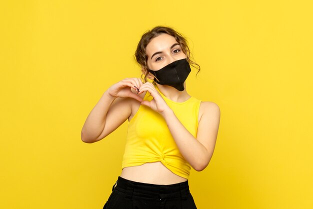 Front view of young woman in mask sending love on yellow wall