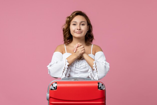 Front view of young woman posing and smiling with red vacation bag on pink wall