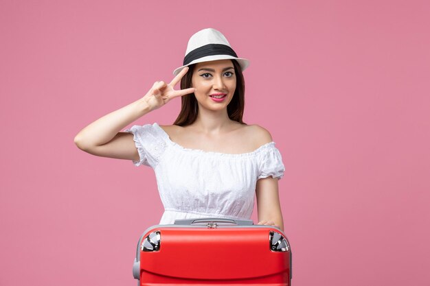 ピンクの壁の休暇の飛行機の航海夏の感情旅行で赤い休暇のバッグでポーズをとる正面図の若い女性