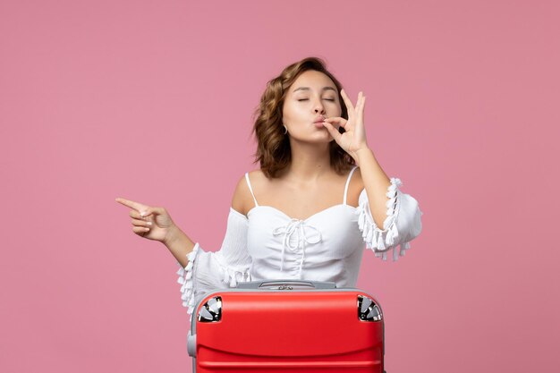 Front view of young woman posing with red vacation bag on pink wall