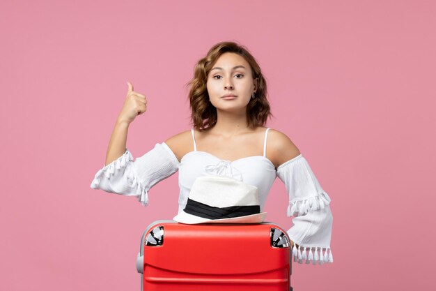 Front view of young woman preparing for vacation with red bag posing on pink wall