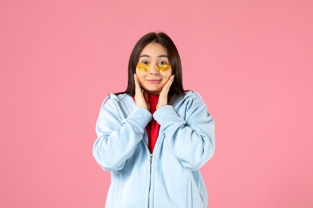 front view of young woman putting eye patches under her eyes on pink wall