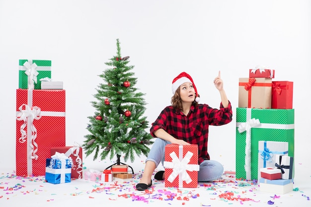 흰 벽에 크리스마스 선물과 작은 크리스마스 트리 주위에 앉아있는 젊은 여자의 전면보기