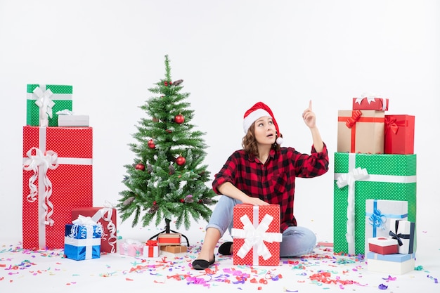 Front view of young woman sitting around christmas presents and little holiday tree on white wall