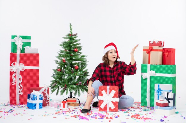 Front view of young woman sitting around christmas presents and little holiday tree on white wall