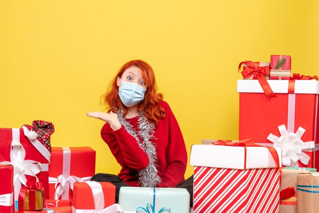 Front view of young woman sitting around christmas presents in mask on yellow wall