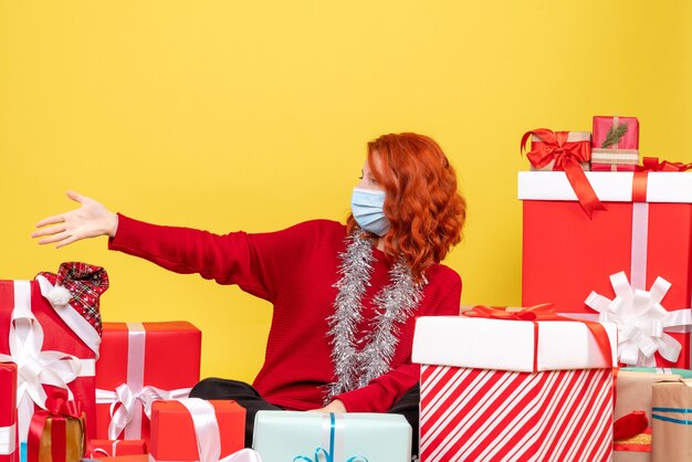 Front view of young woman sitting around christmas presents in mask on yellow wall