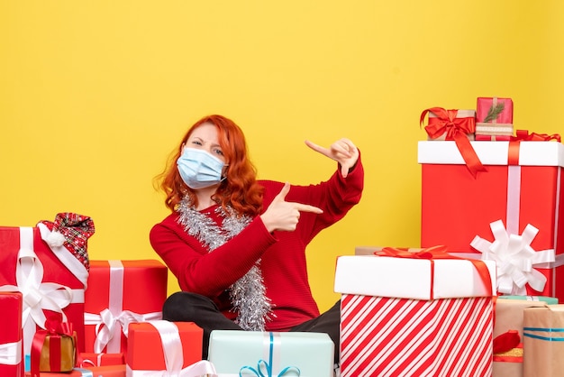 Front view of young woman sitting around christmas presents in mask on yellow wall