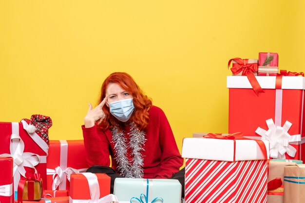 Front view of young woman sitting around christmas presents in mask on yellow wall