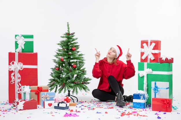 Front view of young woman sitting around different holiday presents on white wall