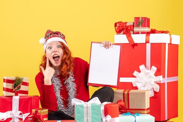 Front view of young woman sitting around different xmas presents on a yellow wall