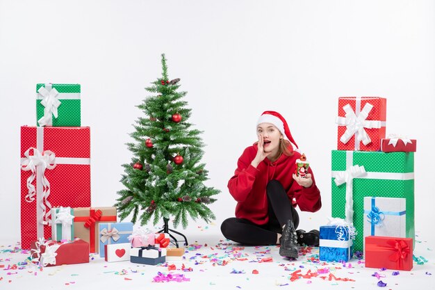 Front view of young woman sitting around holiday presents holding something on white wall