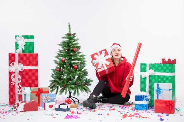 Front view of young woman sitting around holiday presents on a white wall