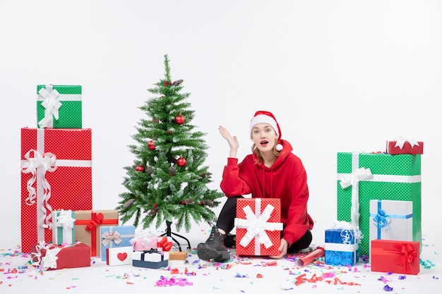 Front view of young woman sitting around holiday presents on white wall