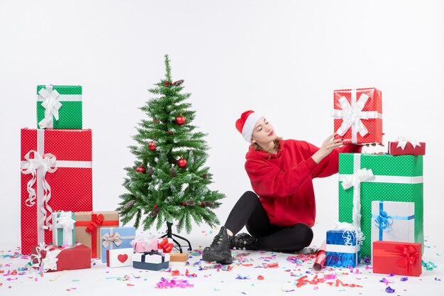 Front view of young woman sitting around holiday presents on white wall