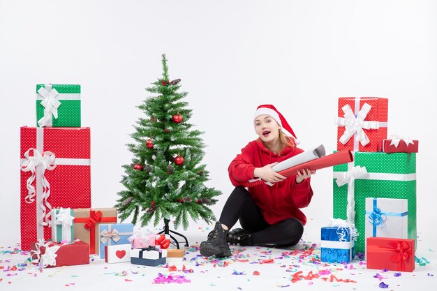 Front view of young woman sitting around holiday presents on white wall